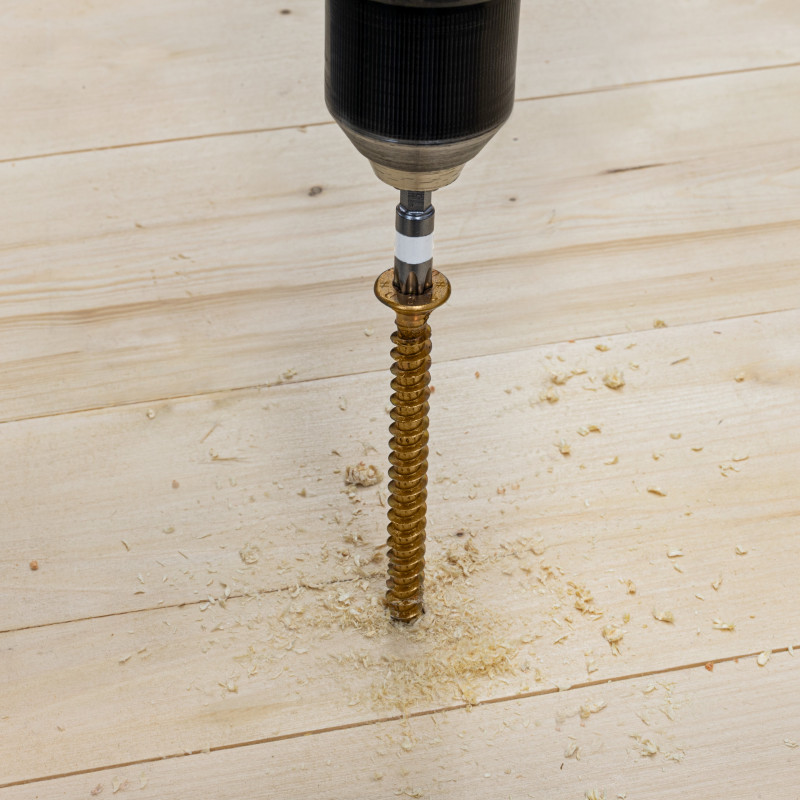 Der Kopf eines motorisierten Schraubers dreht eine gold- oder bronzefarbene Schraube in eine hölzerne Fläche ein. Holzmehl!